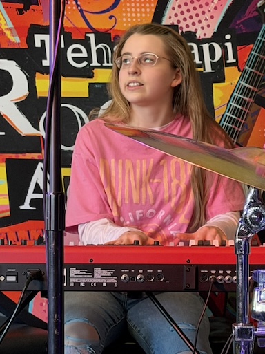 Eliza playing a keyboard with a banner of the Tehachapi Rock Academy logo behind her